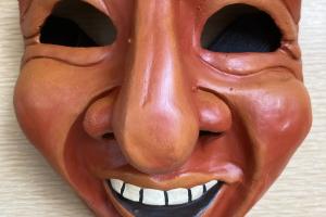 Earth-colored cartoonish laughing mask, with a big nose and empty eye sockets