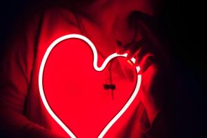 A person holding a neon heart close to their torso, bathed in red light from the heart