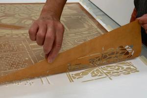 a pair of hands gently remove a thin stencil from a piece of fabric on a table