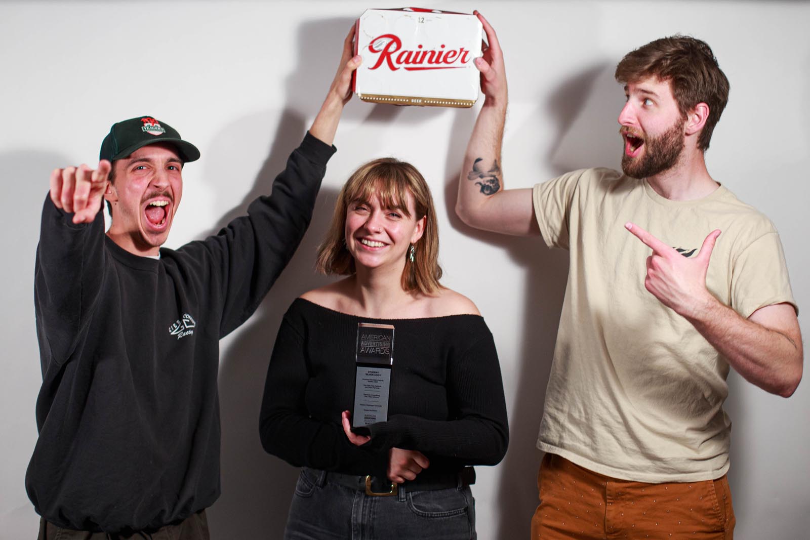 two excited students hold a box of Rainier beer over a third student who holds a trophy