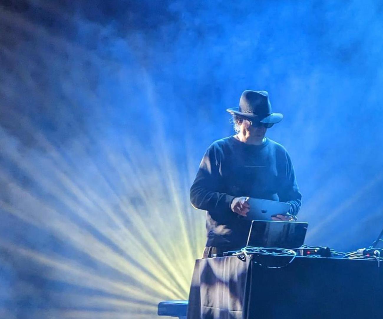 Carl Stone holding a laptop above a laptop at a DJ table surrounded by laser lights