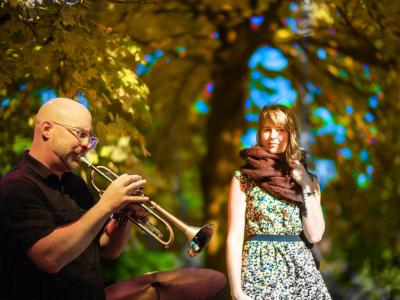 A bald man with glasses plays trumpet under colorful trees while a woman in a floral dress and scarf listens, enjoying the autumn ambiance.