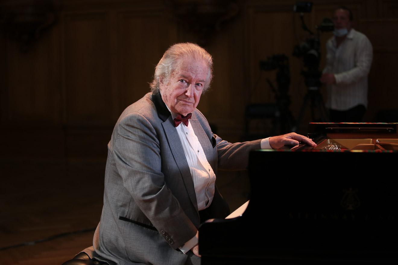 Mikhail Voskresensky in a gray suit, sits at a piano with one hand on top, and turns slightly to look at the camera