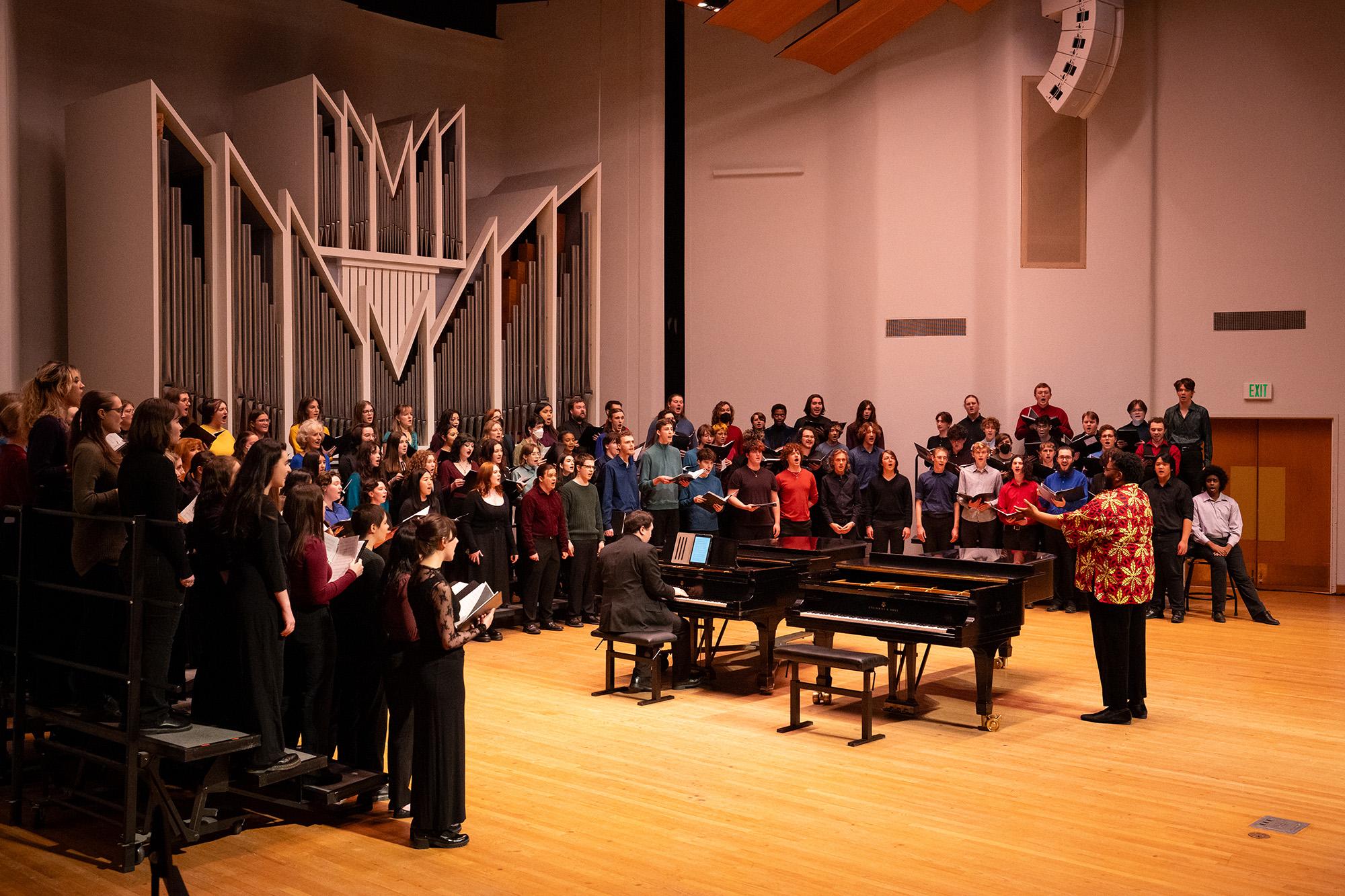 A conductor in front of two pianos - one being played - and surrounded by a singing choir in Western's concert hall