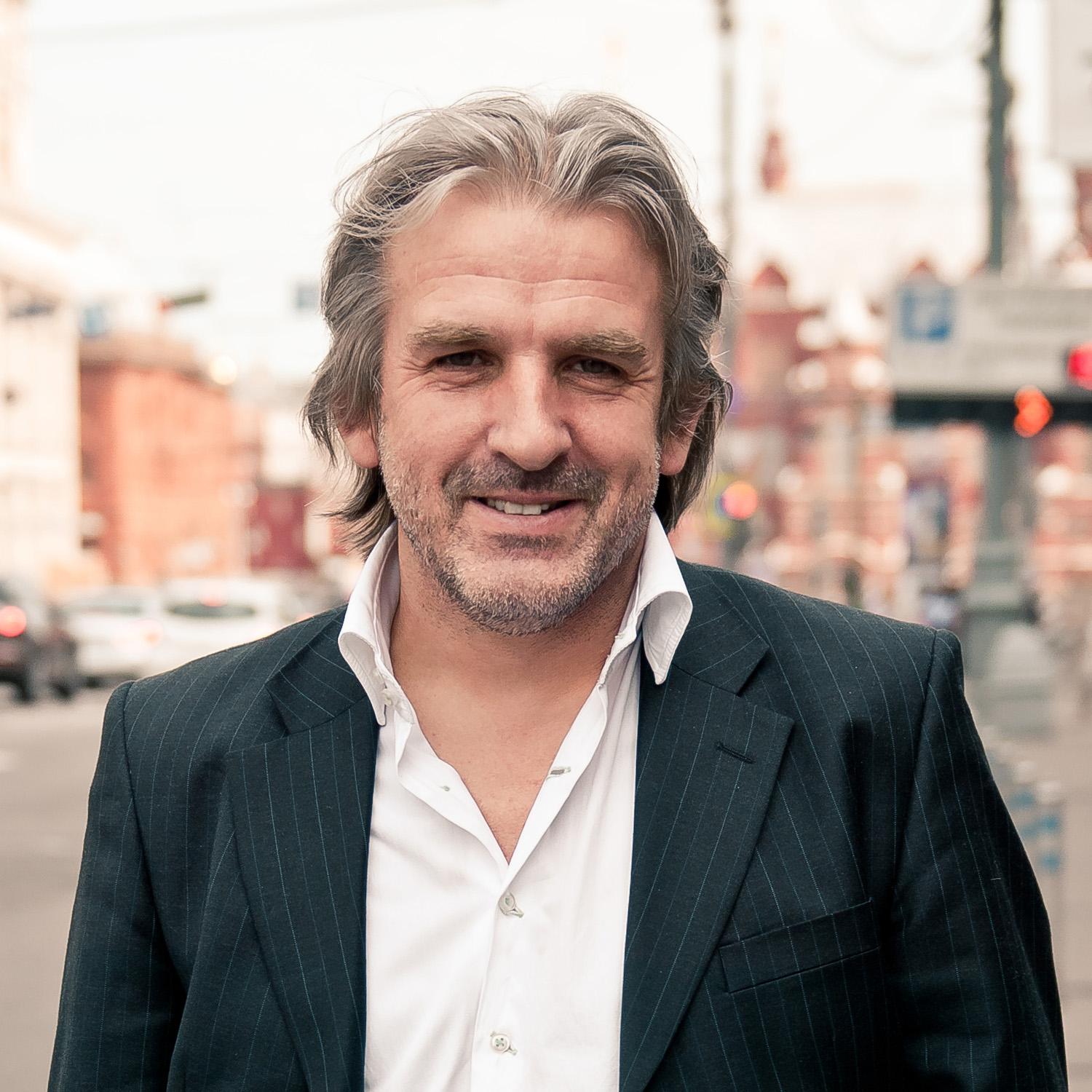 Barry Douglas in formal business wear, smiling and squinting slightly in front of a blurry street scene