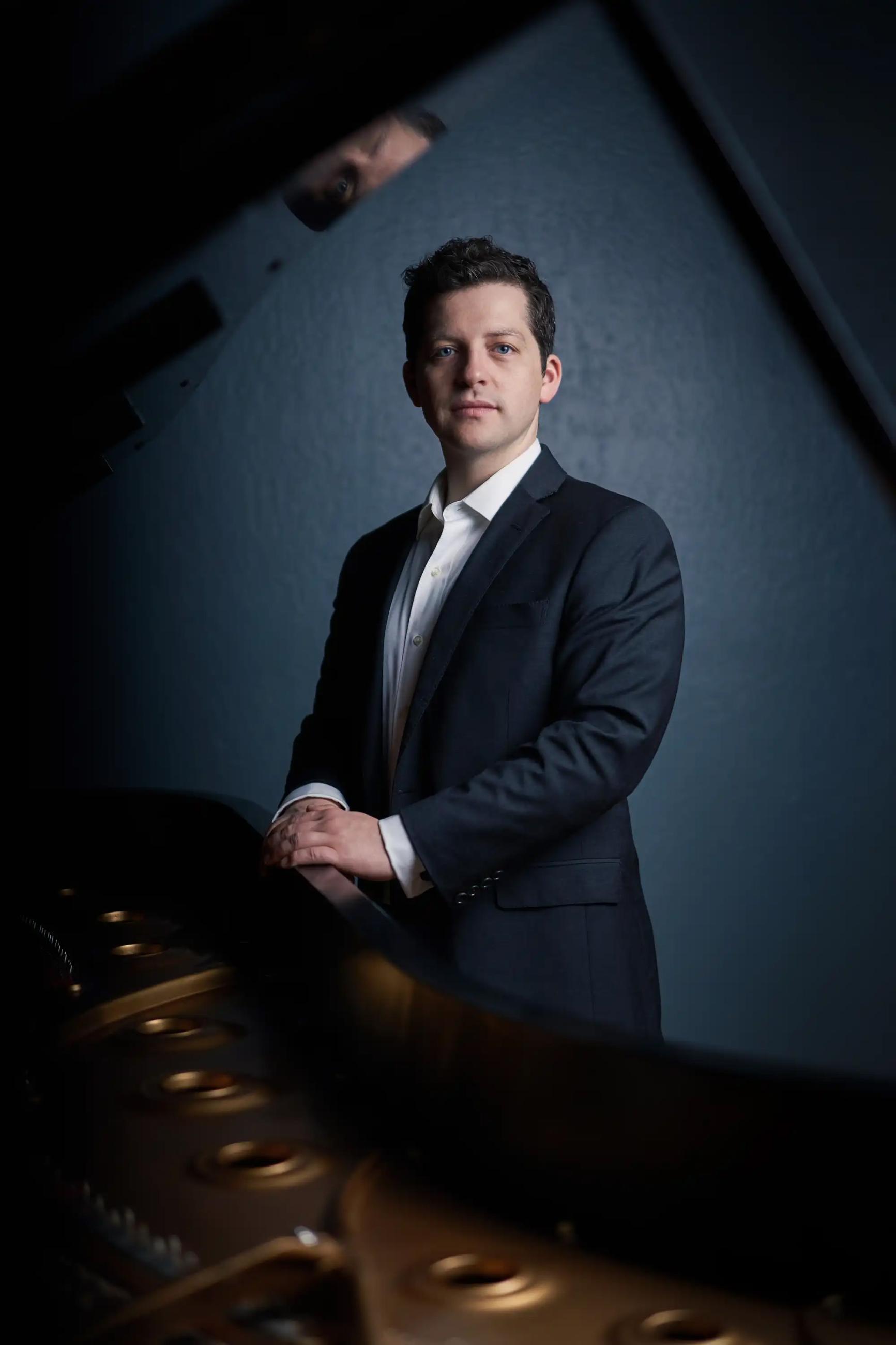 Henry Kramer standing spotlit in a suit with perfect posture, a calm face, and hands resting on top of a piano