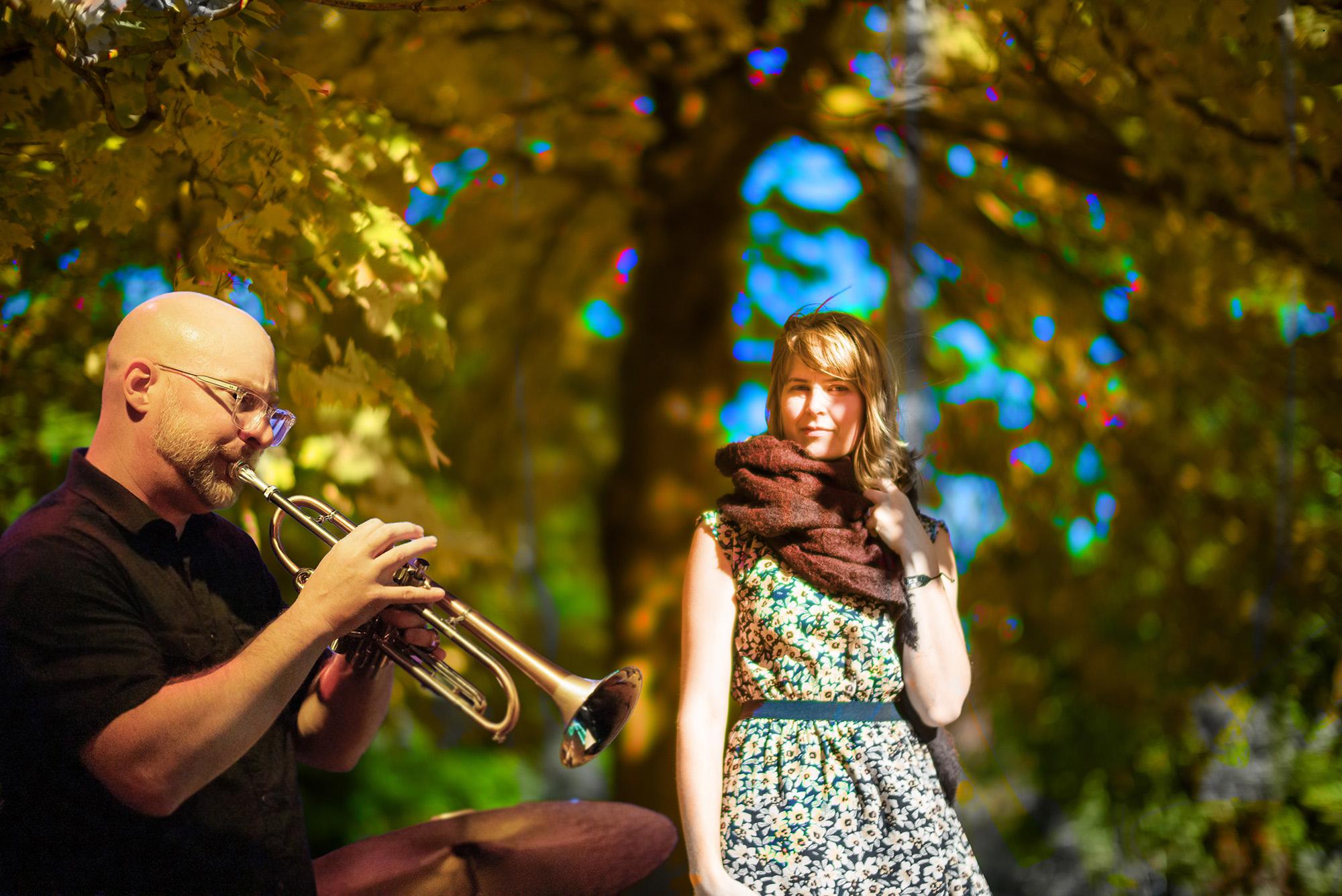 A bald man with glasses plays trumpet under colorful trees while a woman in a floral dress and scarf listens, enjoying the autumn ambiance.