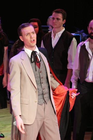singing actor in suit, waistcoat, and string tie. Other actors in vests look on.