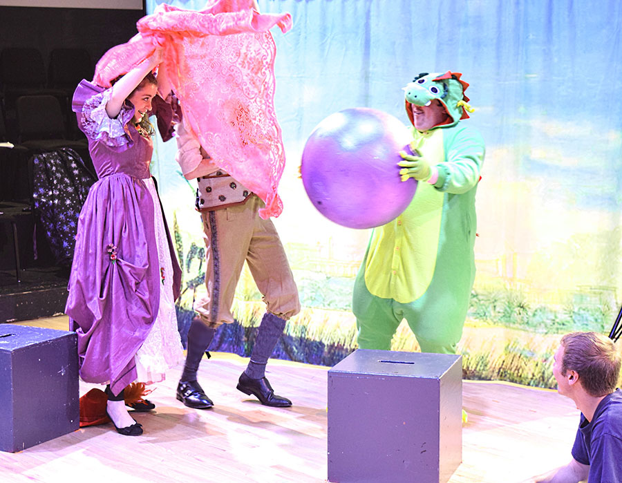 two brightly clothed actors flail about near person in dragon suit holding purple ball.