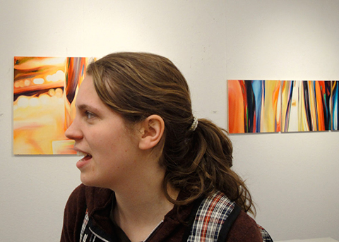 A female with a ponytail in front of two paintings on the wall looks off-camera with an open mouth 