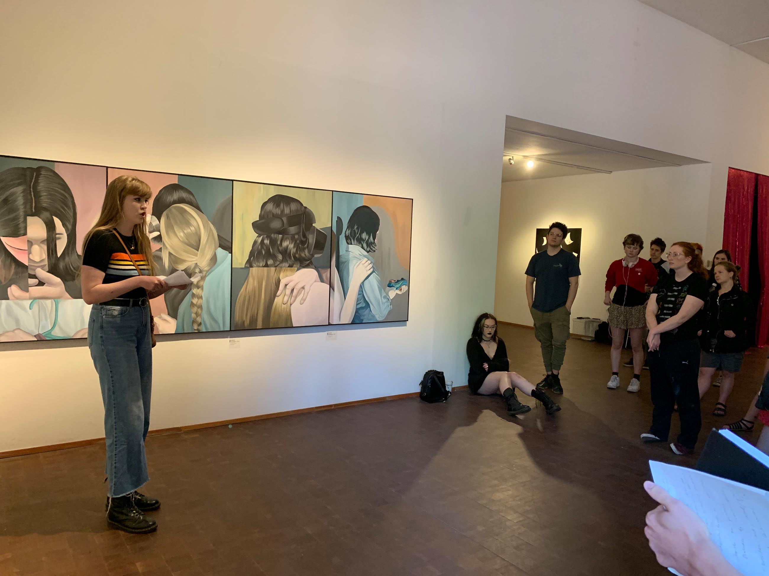a painting student stands in front of a painting on a wall and speaks to a small group