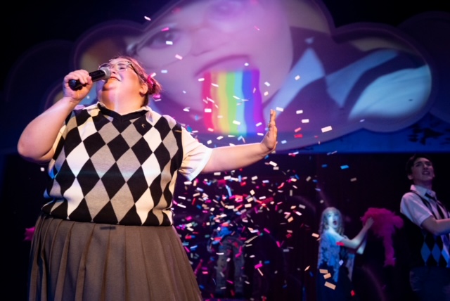 A person singing expressively into a microphone. Behind them confetti falls in front of a projection of a face spitting a rainbow.
