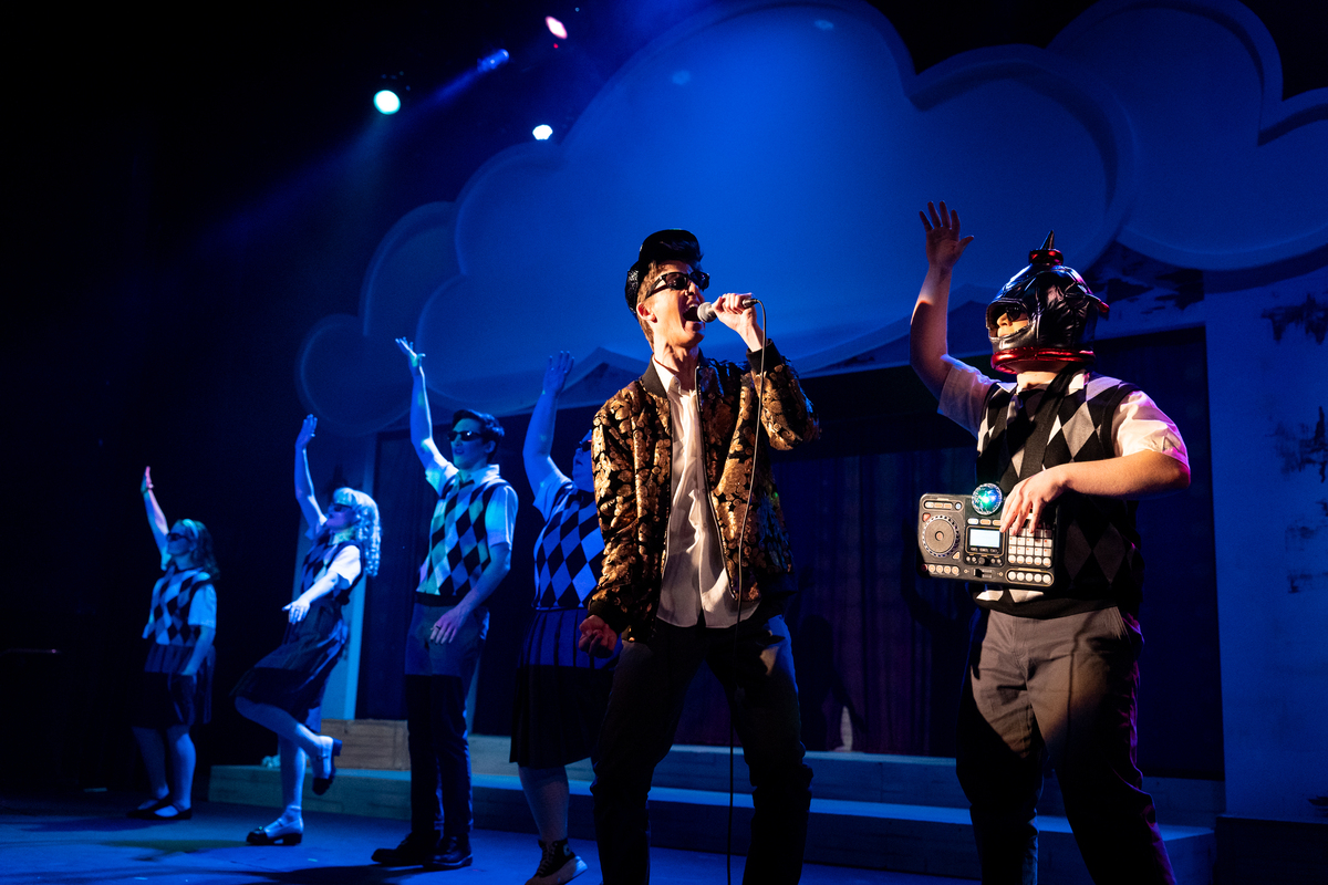 A person in a funky outfit sings loudly into a microphone in front of a line of dancers. A person wearing a helmet and ghetto blaster dances nearby.