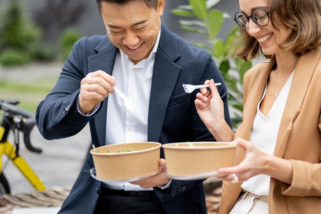 Joyful asian businessman and caucasian businesswoman eating food and talking on an outdoor bench