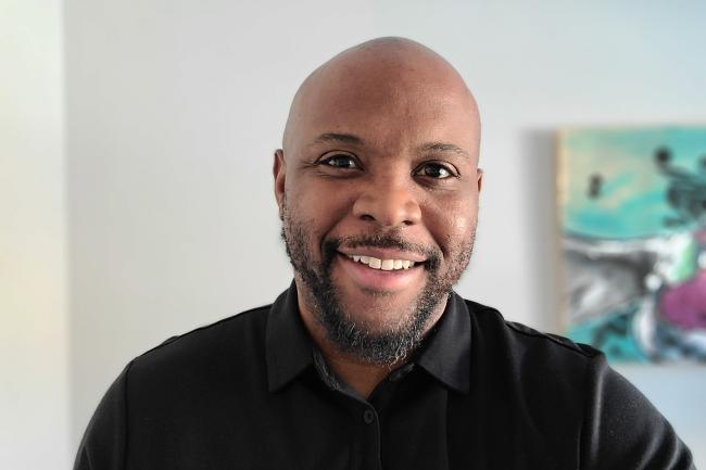Idris Goodwin sporting a short beard and a friendly smile, in front of a blank wall with a blurred painting