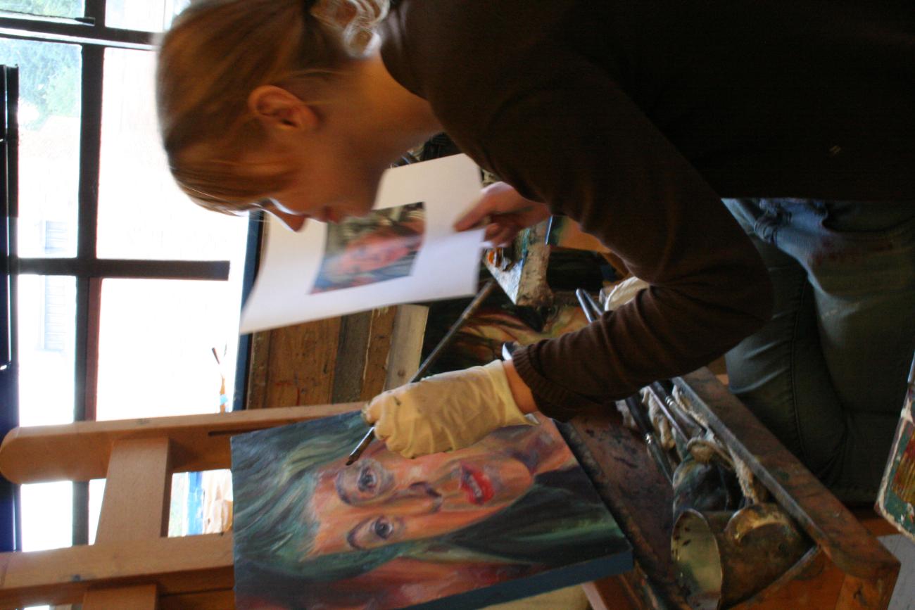 A student smiles as they add to a painting on an easel, holding a portrait photo in one hand which they have based the painting on