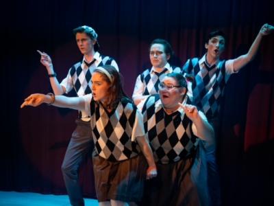 Four actors in checkered shirts grouped together, pointing outward accusationally