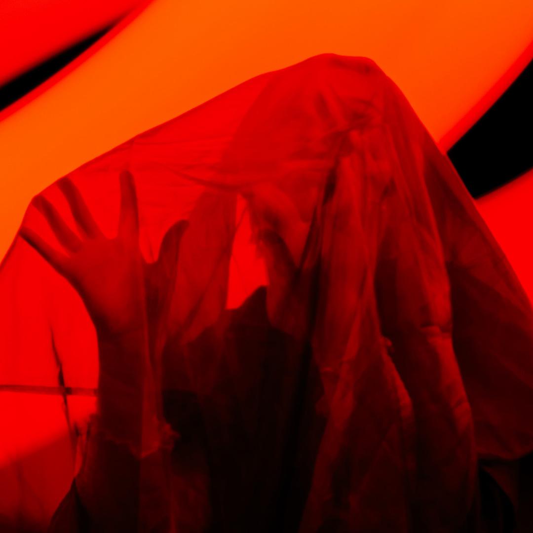 saturated red and orange light illuminate a person under a veil with one held up, palm out, fingers open.