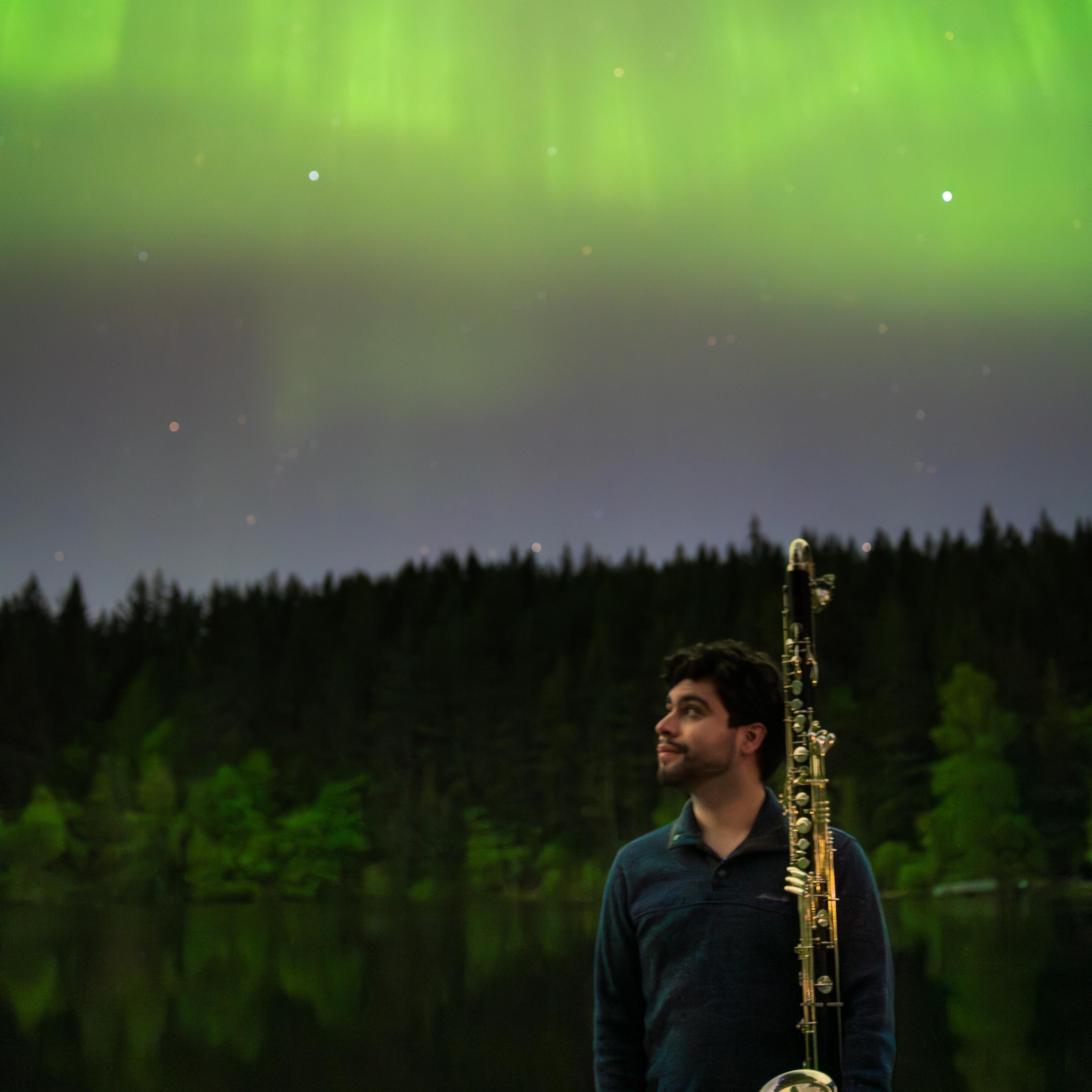 David Bissell standing holding a clarinet, smiling up at a sky filled with aurora borealis